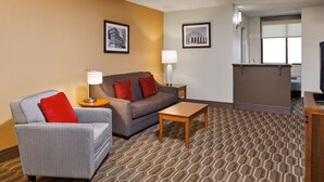 A hotel room with a grey sofa, a small table, a lamp, and a kitchen area.