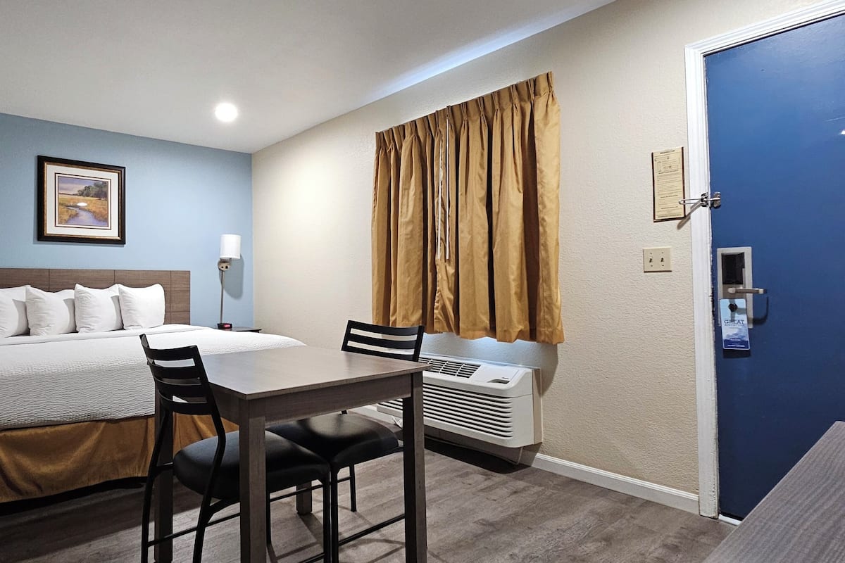 A hotel room with a bed, a desk with chairs, a standing lamp, an air conditioner, and a door with a blue panel.