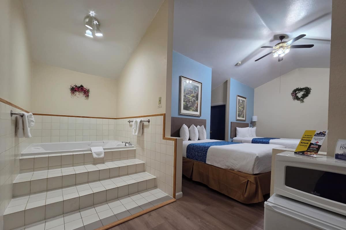 A hotel room with a bathtub, a bed with a blue bedspread, a ceiling fan, and a refrigerator.
