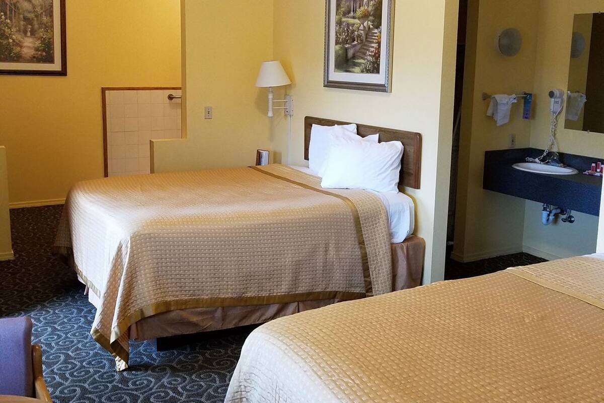A hotel room with two beds, a desk, a chair, a lamp, a painting, and a bathroom with a sink and mirror.