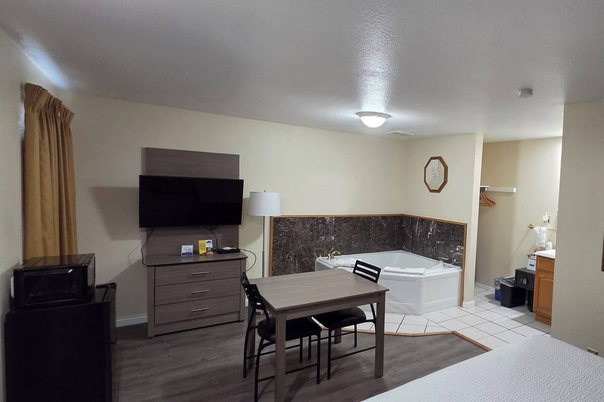 A hotel room with a flat-screen TV, a desk with a chair, a bathtub, a framed picture on the wall, and a small trash bin.
