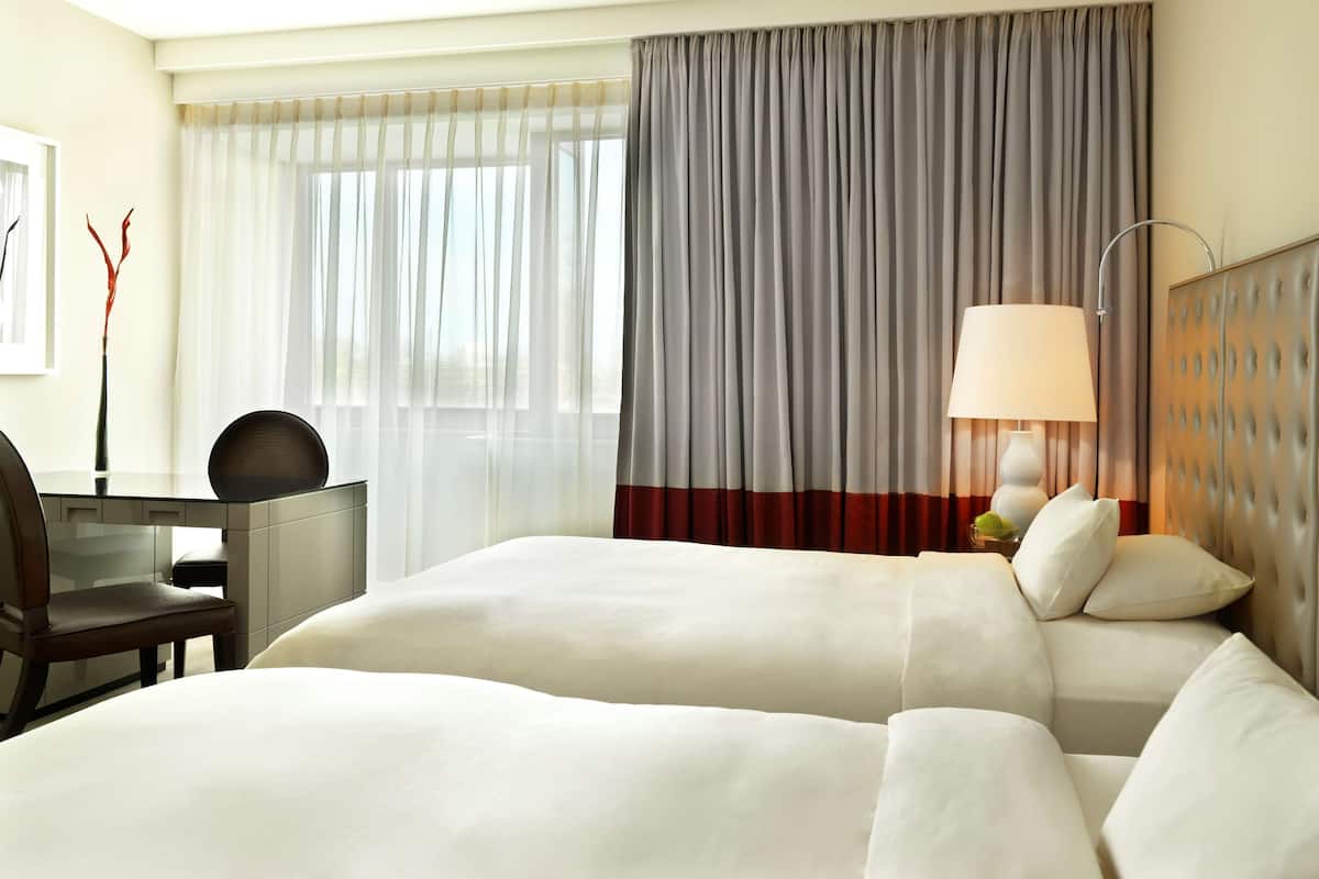 A hotel room with two beds, a desk, a chair, a lamp, and a view of the outdoors.