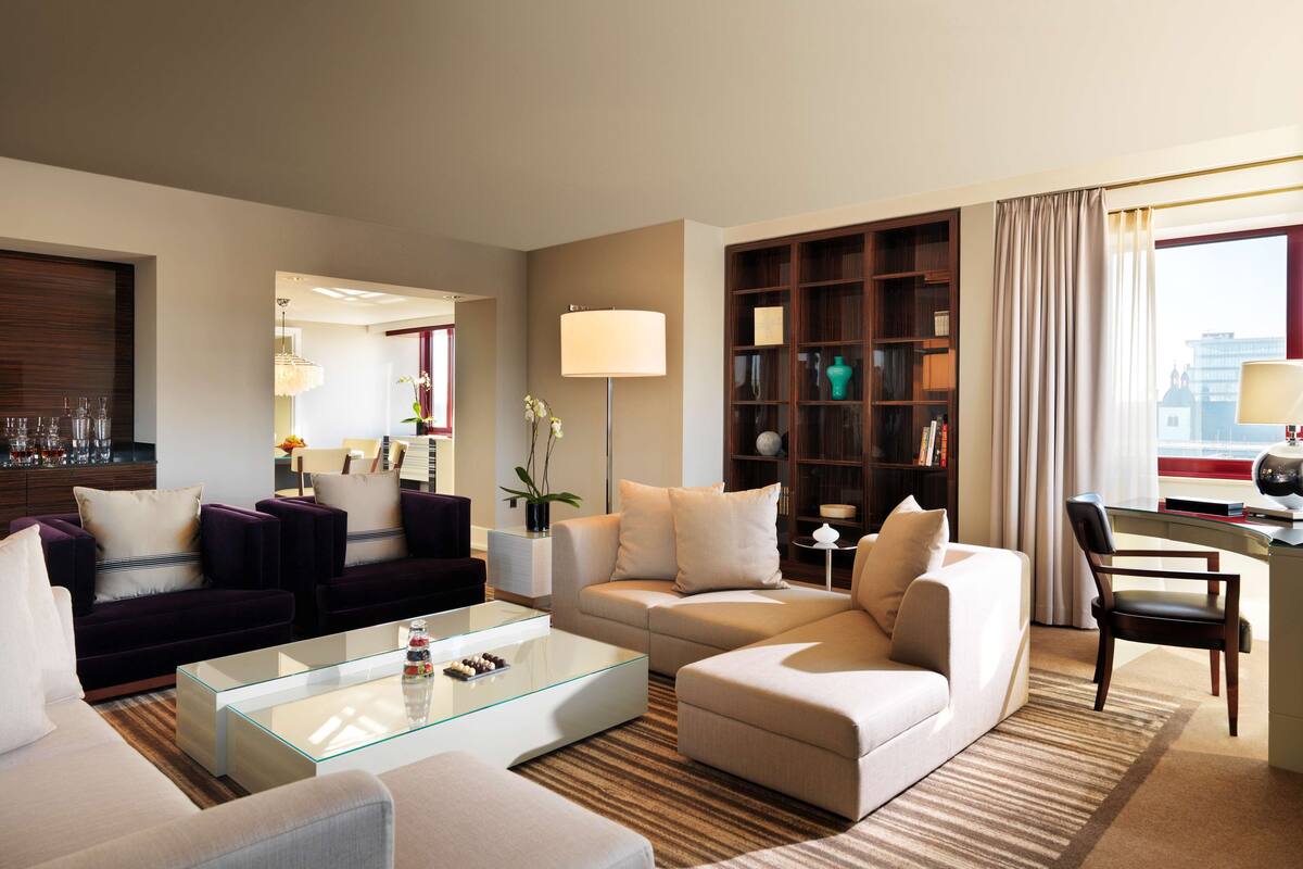 A modern living room with a glass coffee table, two sofas, a bookshelf, and a view of a cityscape through a large window.