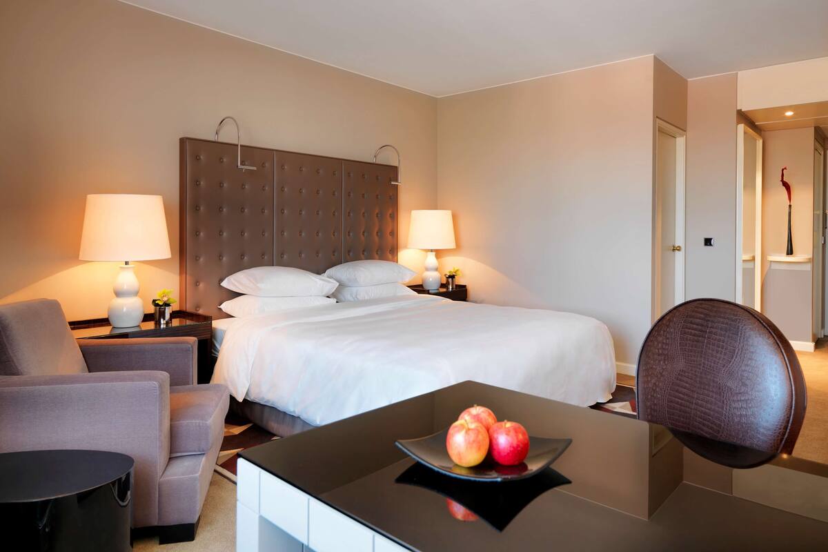 A hotel room with a bed, two bedside tables with lamps, a chair, a desk with a plate of apples, and a door with a red sculpture.