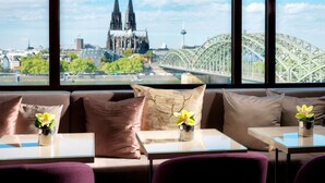 Ein Hotelzimmer mit großem Fenster und Blick auf ein Dom und eine Brücke. Zwei Tische mit Blumenvasen und eine Couch mit Kissen sind vorhanden.