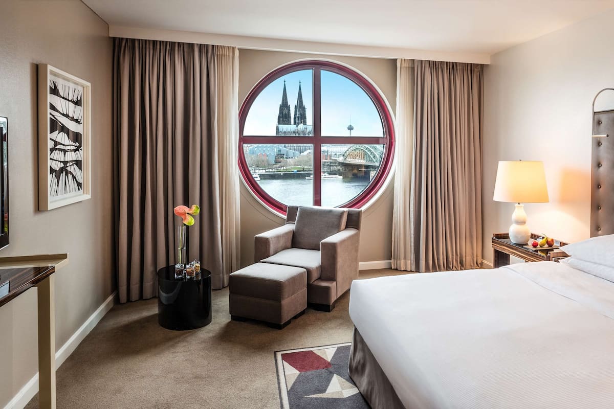 A hotel room with a large bed, a round window with a view of a cathedral and a bridge, a chair, a TV, and a lamp.