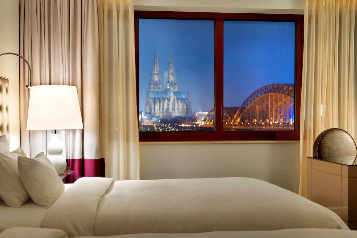 A hotel room with a large bed, a nightstand with a lamp, and a view of a cityscape including a cathedral and a bridge.