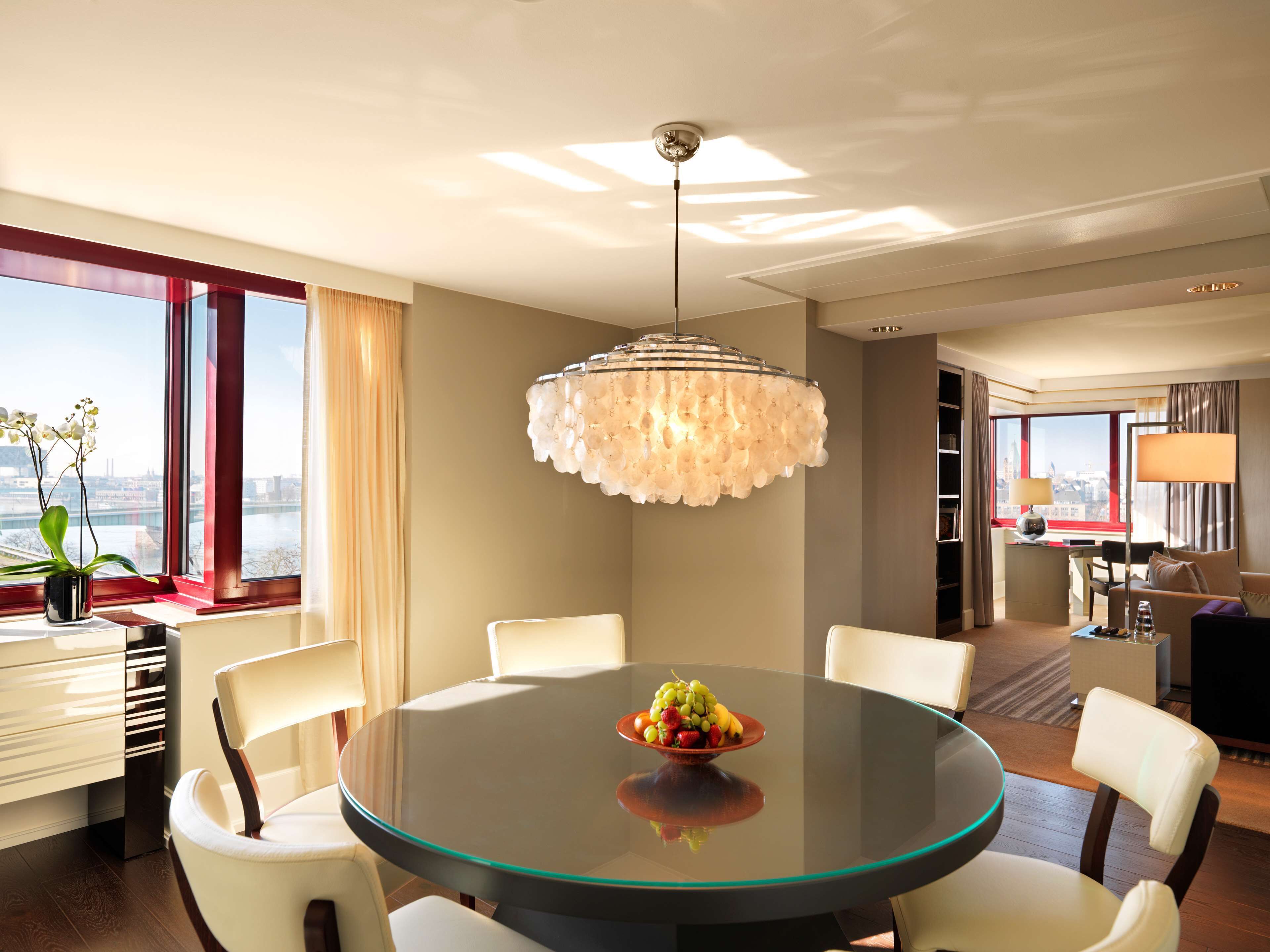 Une chambre d’hôtel moderne avec une table ronde en verre, des chaises blanches, un lustre et une vue sur la ville.