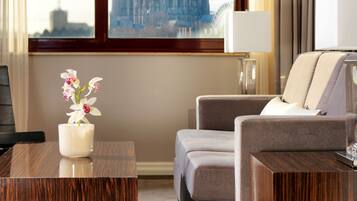 A hotel room with a large window offering a view of a cathedral and a bridge. There is a wooden coffee table with a vase of flowers, a grey sofa, and a lamp on a side table.