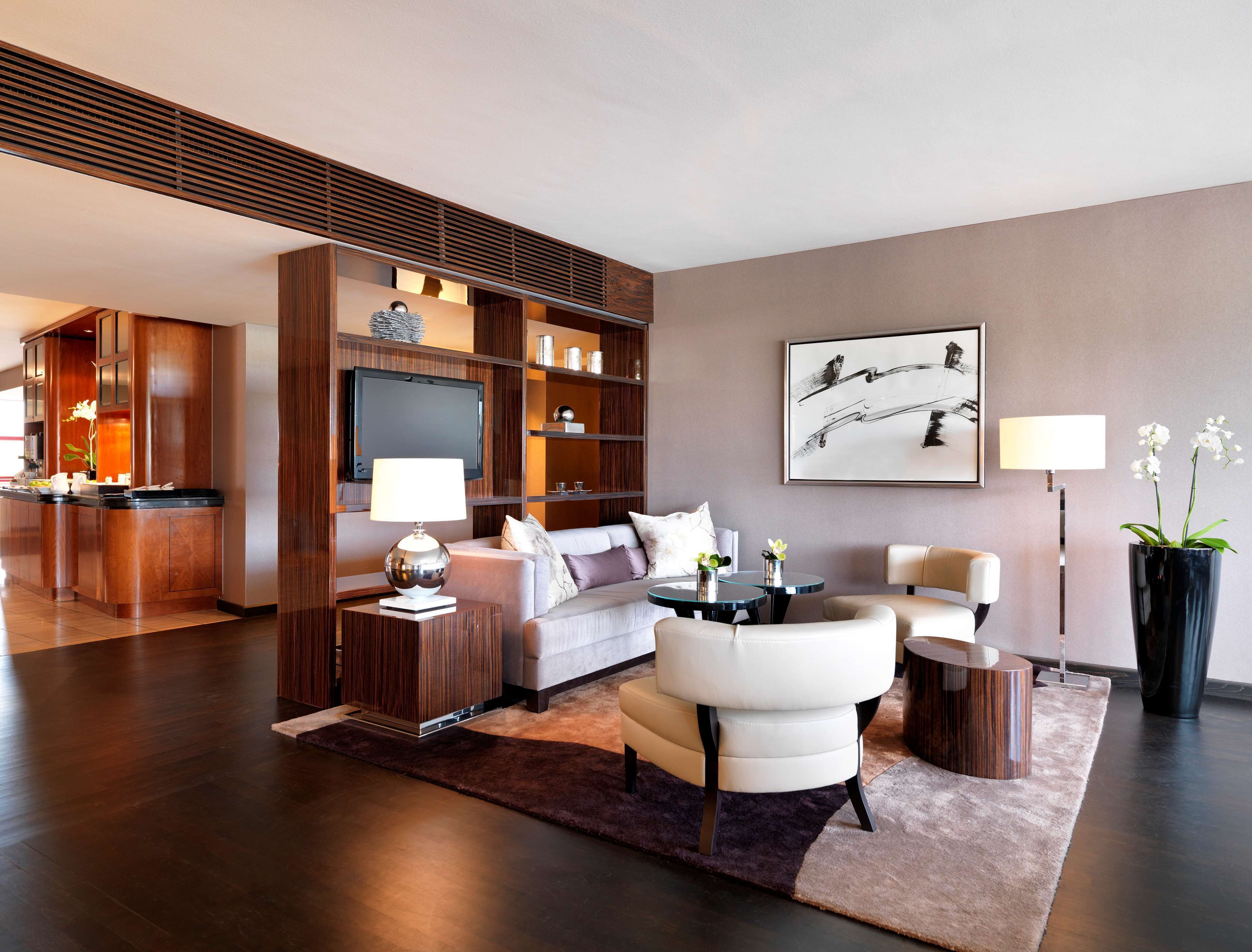 A modern living room with a sofa, armchairs, a coffee table, and a TV. There is a kitchen area with wooden cabinets and a dining table.