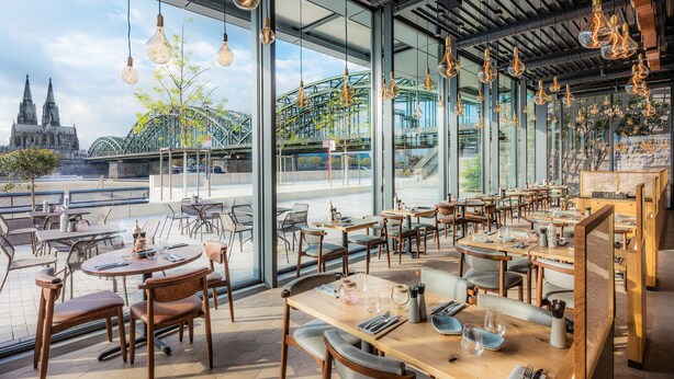 Ein modernes Restaurant mit großen Fenstern, das Blick auf einen historischen Dom und eine Brücke bietet. Tische sind mit Stühlen bestückt, und das Interieur zeichnet sich durch freiliegende Balken und hängende Leuchten aus.