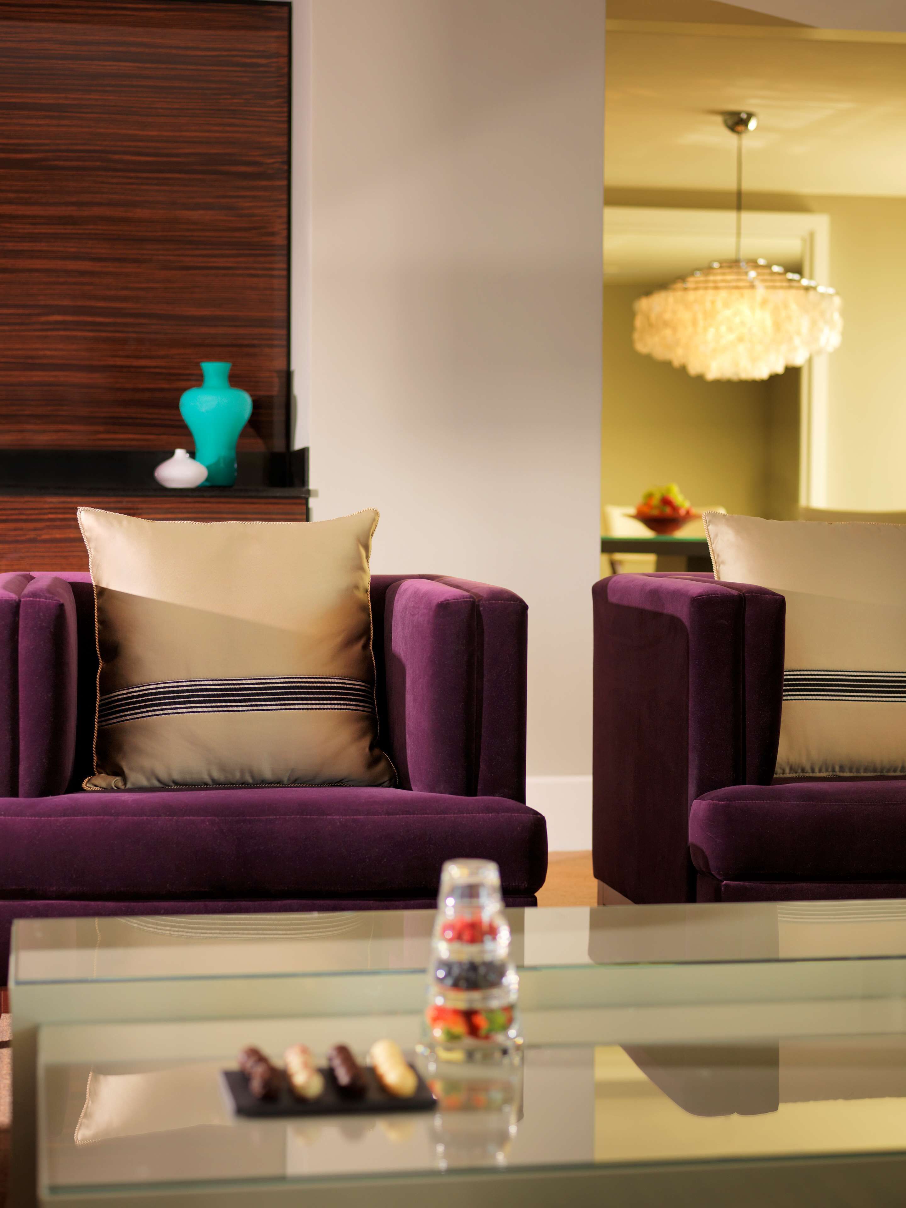 Ein modernes Wohnzimmer mit zwei violetten Sessel, einem Glastisch mit Snacks und einer dekorativen Vase auf einem Holzregal.