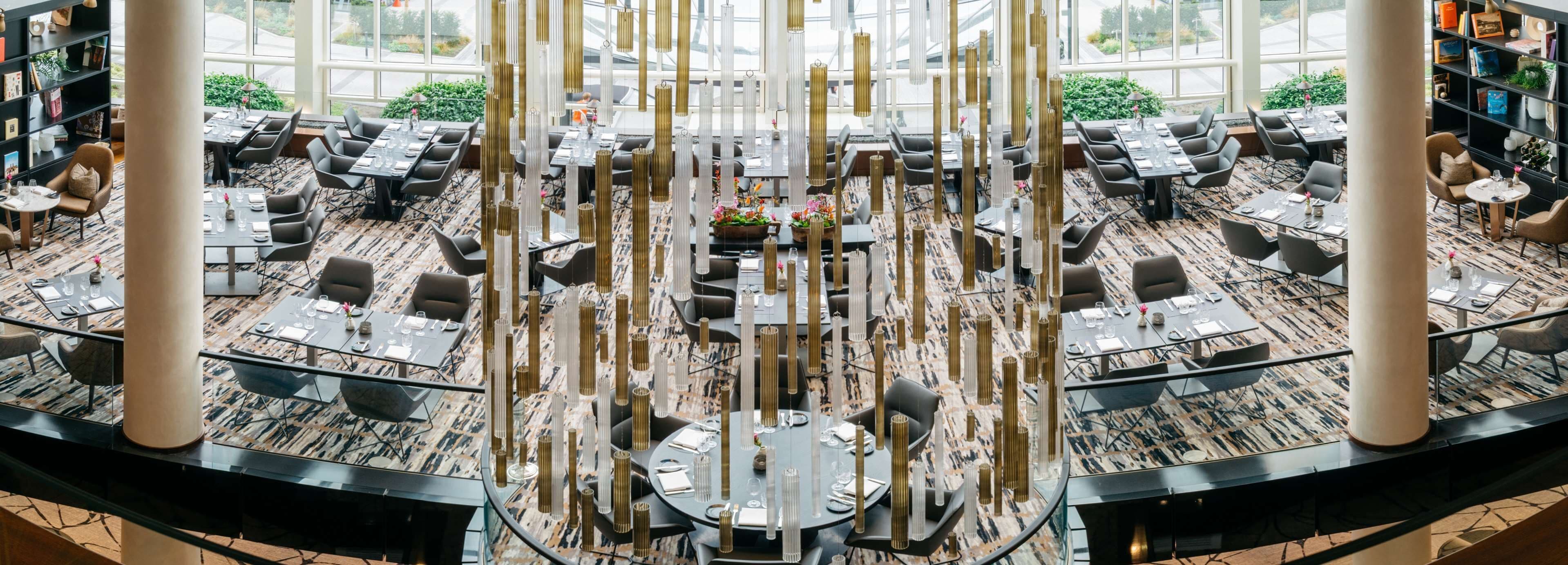 A spacious indoor area with multiple tables and chairs, a central decorative light fixture, and large windows providing natural light.