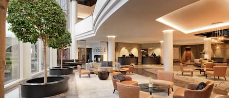 A spacious lobby with a reception desk, seating areas, and a large tree.