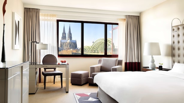 A hotel room with a bed, desk, chair, and a view of a cathedral and bridge.