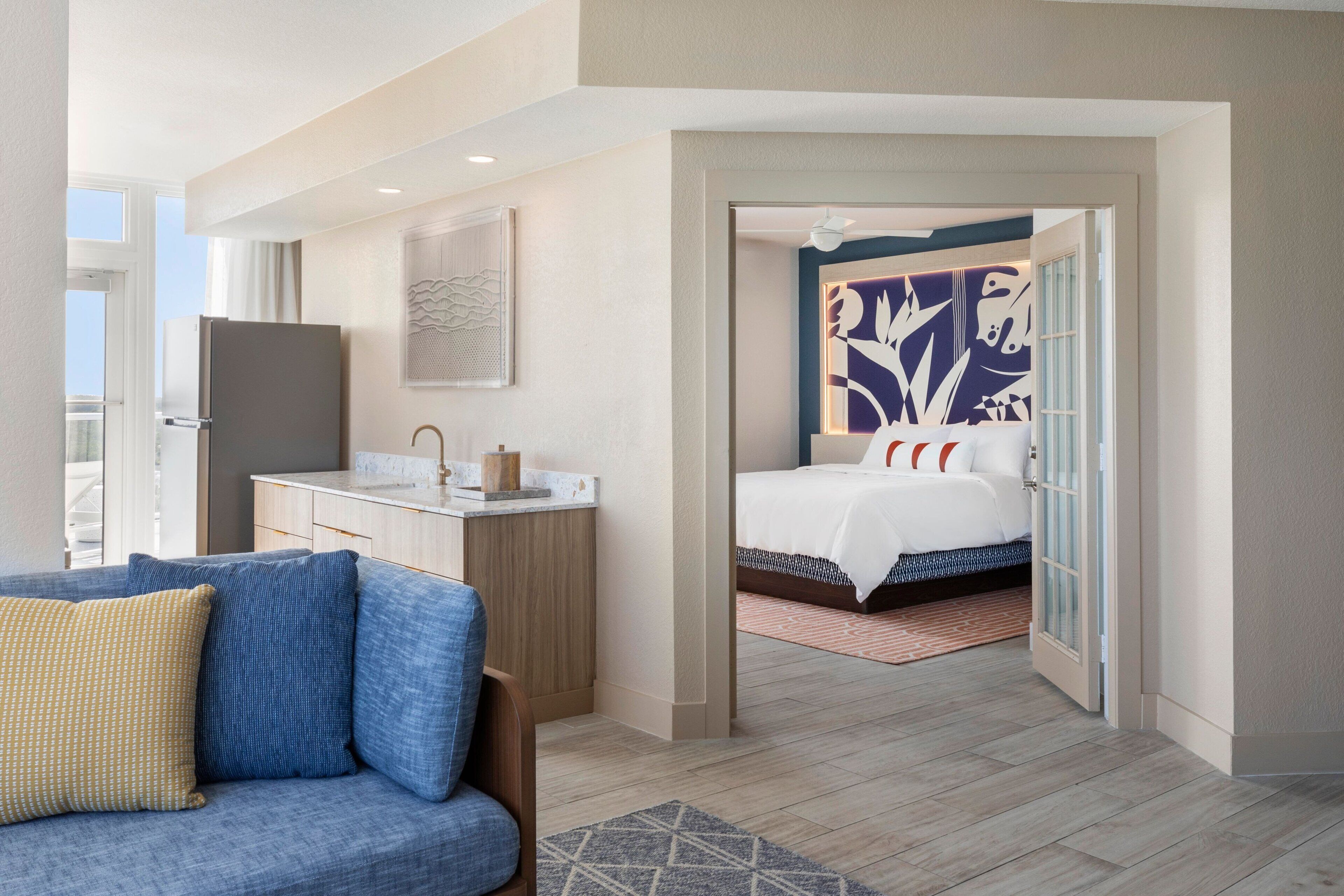 A modern hotel room with a bed, a sitting area with a sofa, a small kitchenette, and a bathroom visible through an open door.
