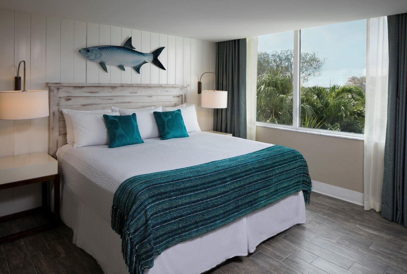 Habitación de hotel con cama, un ventanal con cortinas, un adorno de pez en la pared y una mesita de noche con lámpara.