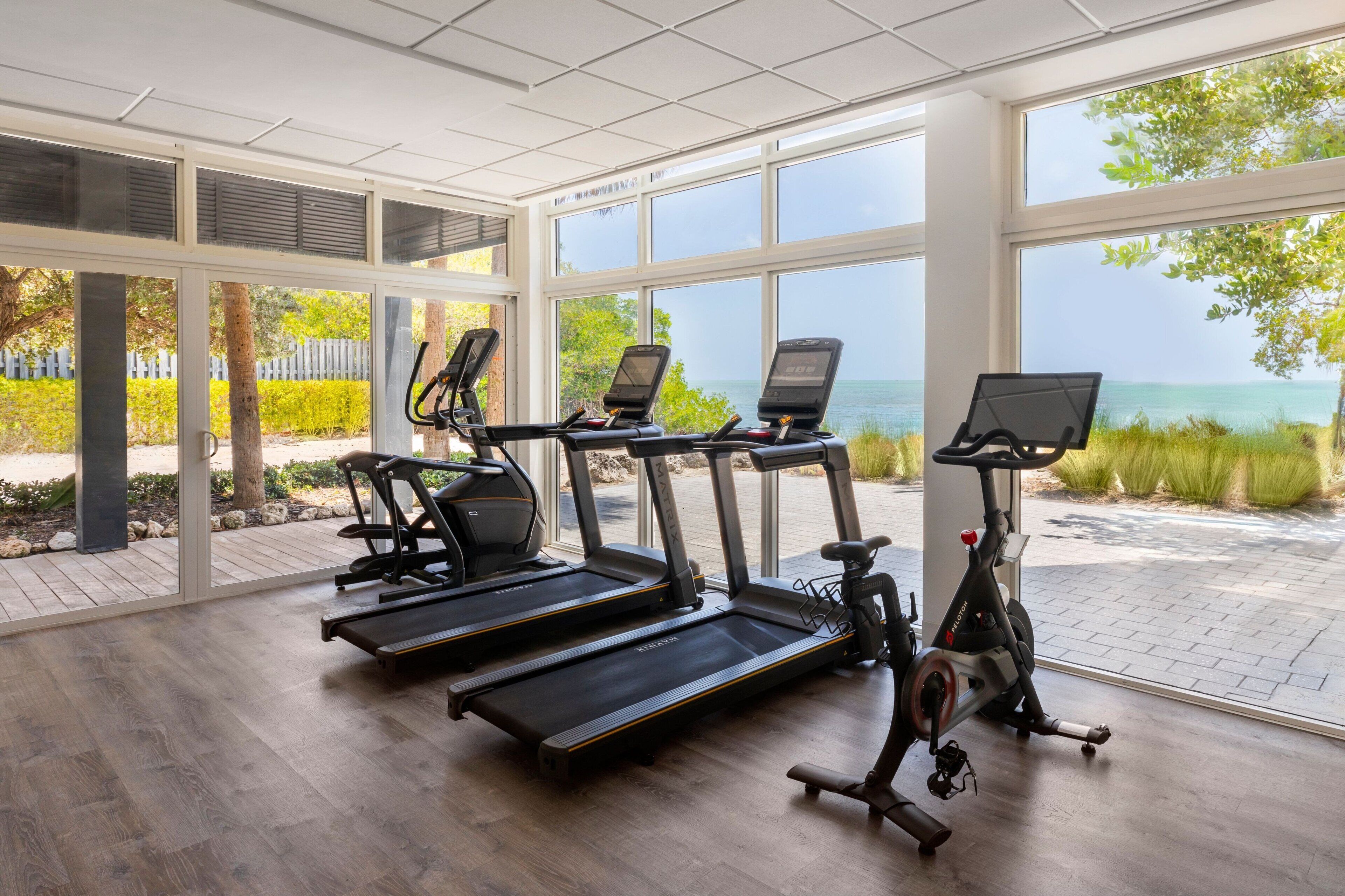 A gym with treadmills, an elliptical, and a stationary bike, large windows with a view of the sea.