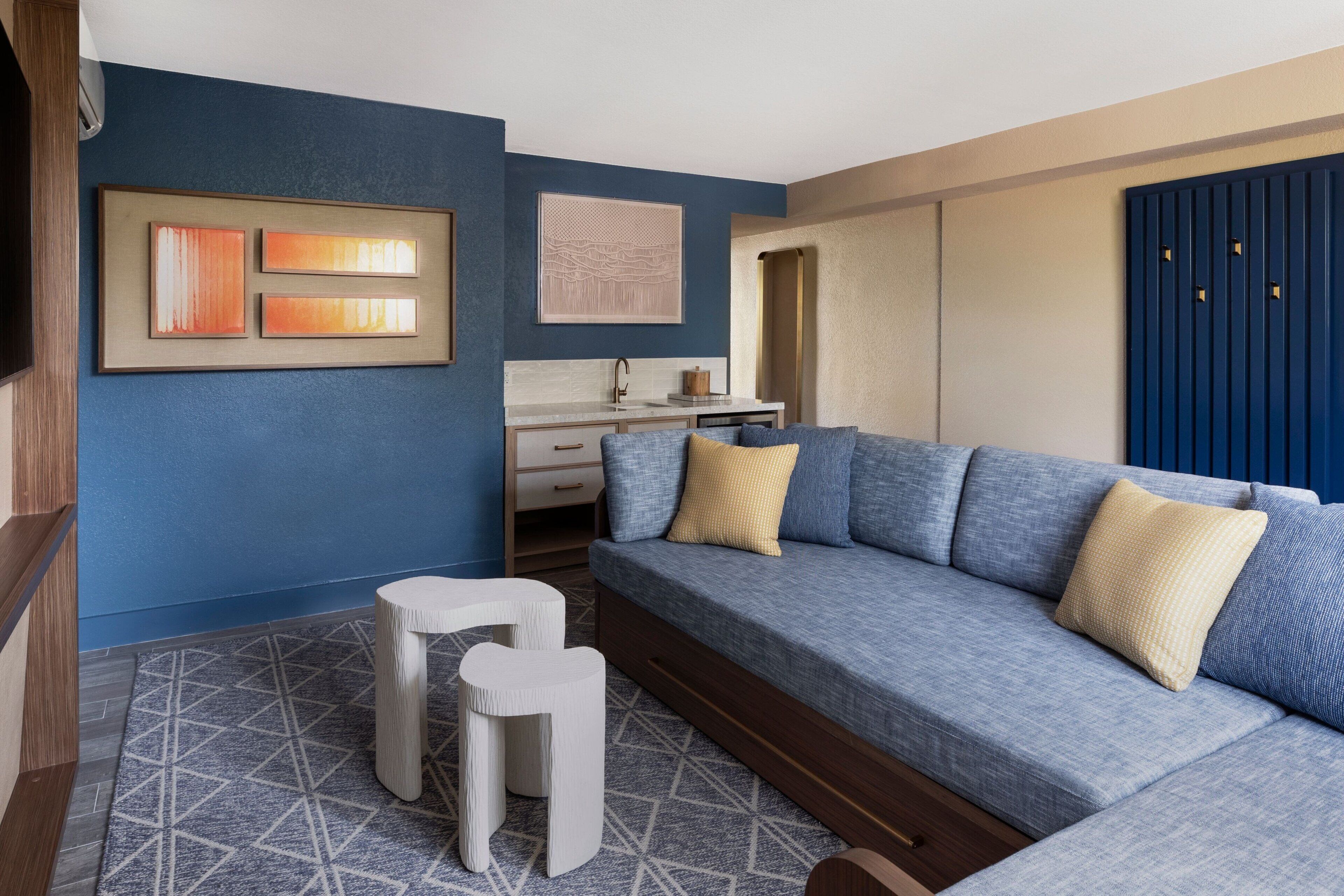 Una sala moderna con un sofá azul, dos taburetes blancos, una zona de cocina con un lavamanos y un armario azul.