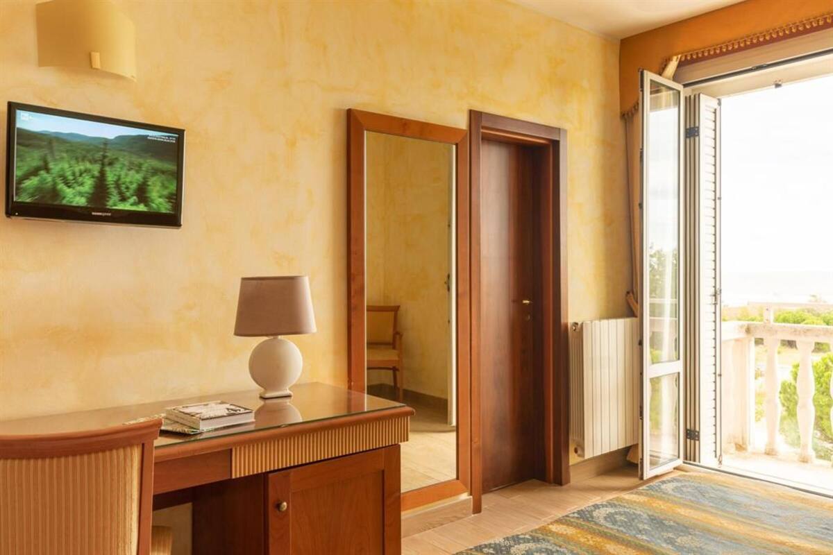A hotel room with a wooden desk, a lamp, a television mounted on the wall, and a sliding glass door leading to an outdoor area.