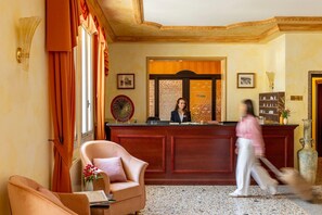 Un'area reception con una scrivania in legno, una receptionist, una poltrona confortevole e un vaso grande.