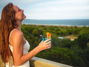 A person holding a drink with a straw and a slice of fruit, overlooking a beach with greenery and the ocean in the background.