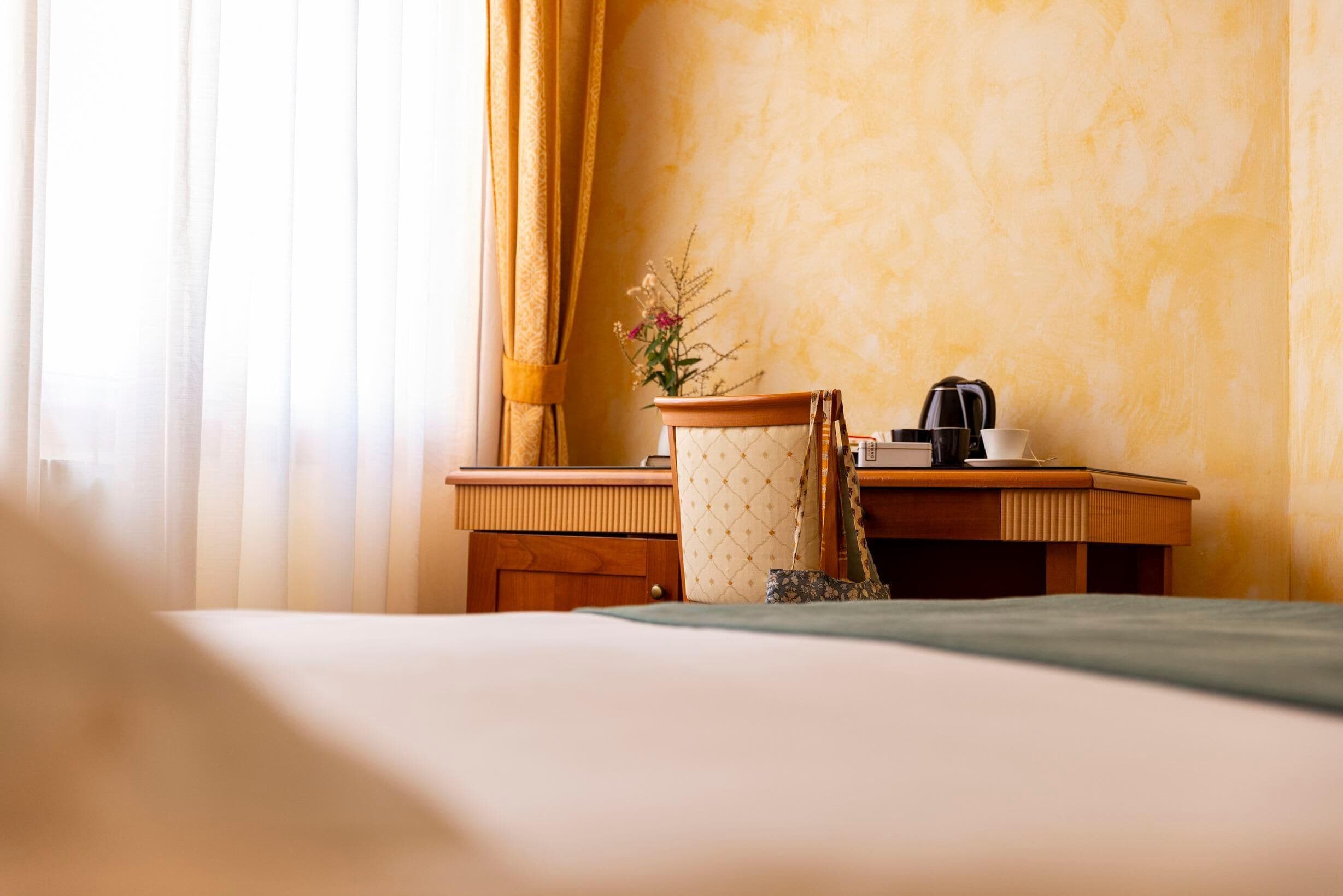 A hotel room with a bed, a wooden desk, a chair, a coffee maker, and a vase with flowers.