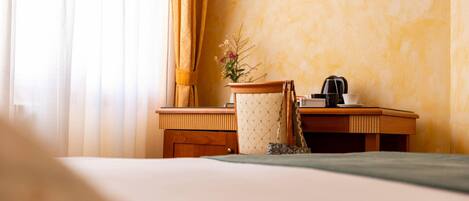 A hotel room with a bed, a wooden desk, a chair, a coffee maker, and a vase with flowers.
