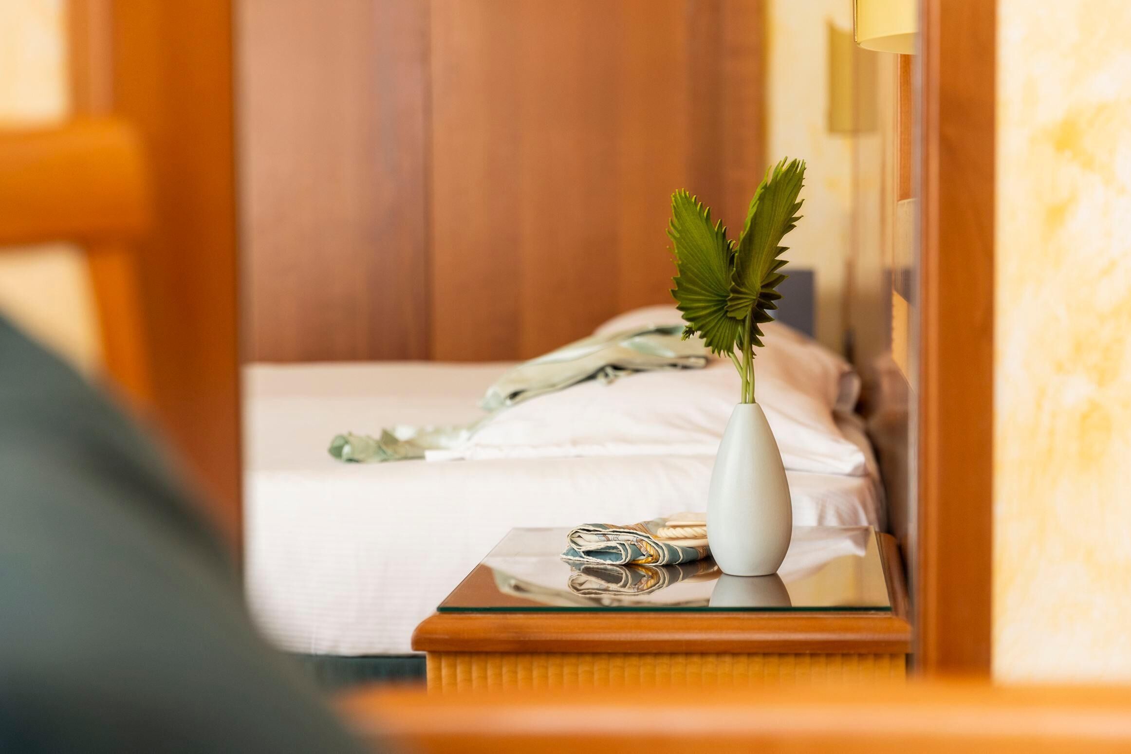 Una camera d'albergo con un letto rifatto, un comodino con un vaso bianco e un paio di occhiali, e una testiera in legno.