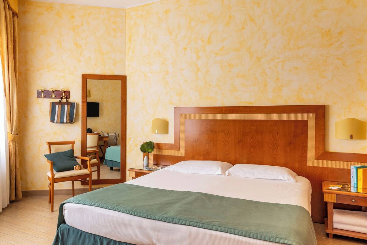 A hotel room with a large bed, a wooden headboard, a desk with a chair, a mirror, and a nightstand with books.