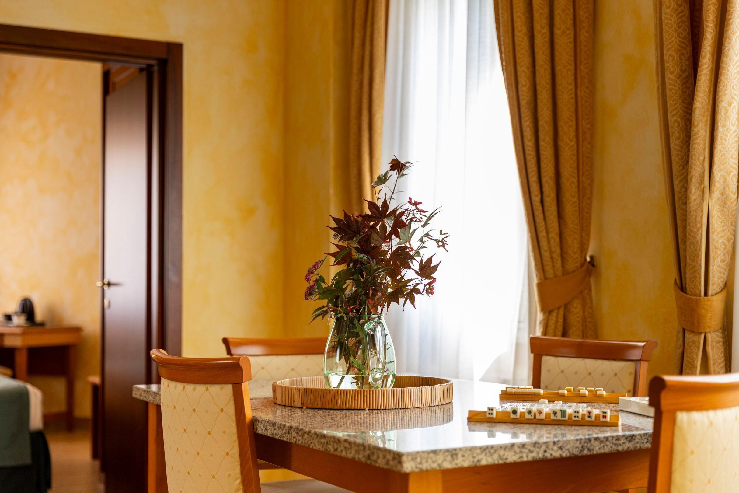 A dining area with a table set for four, a vase with flowers, and a view of a room with a door.