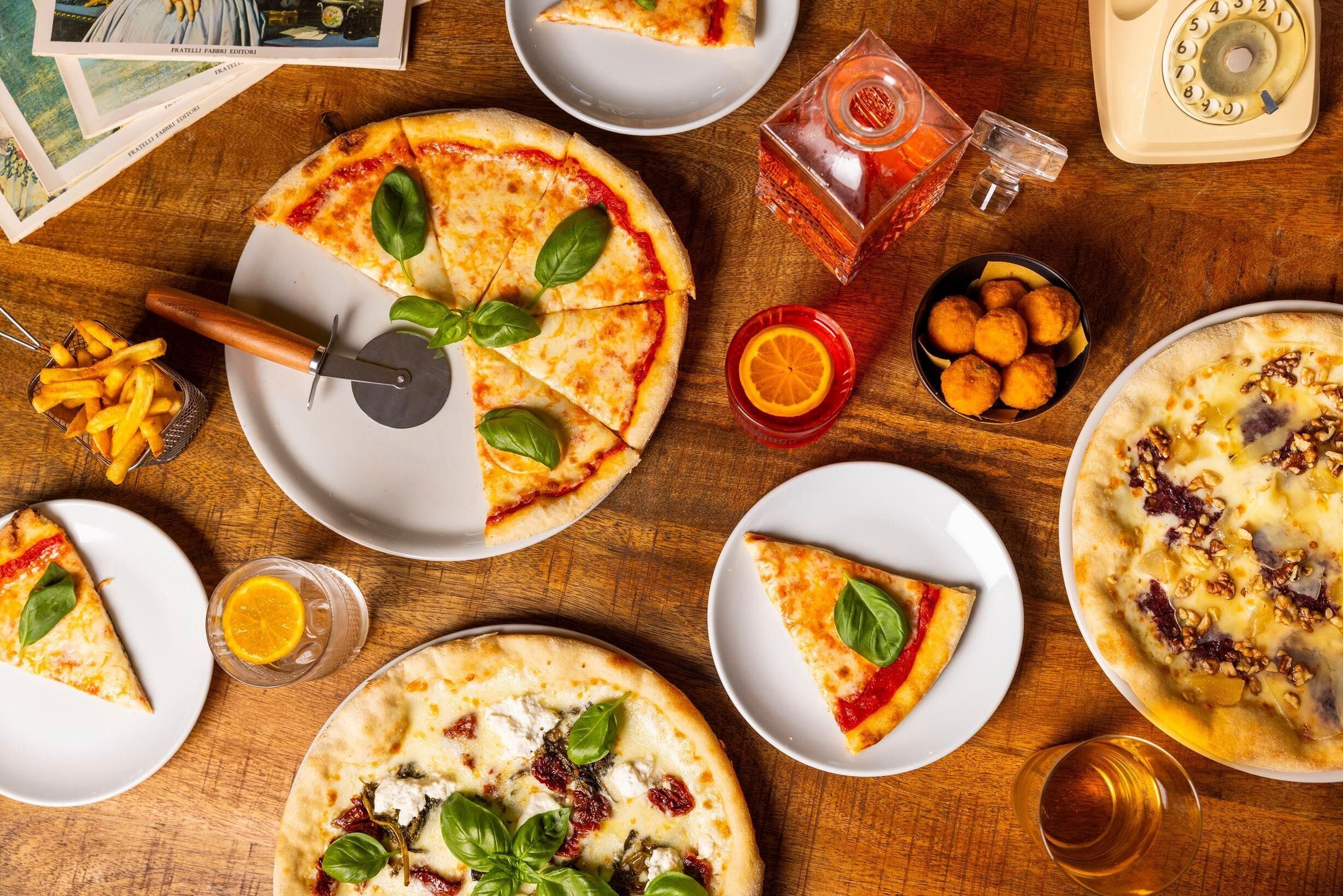 Ein Holztisch mit verschiedenen Pizzen, einem Pizzaroller, einem Glas Orangensaft, einem Glas Bier und einem Salzstreuer.