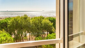 Blick aus einem Fenster auf einen Strand, im Vordergrund Bäume und ein klarer Himmel.