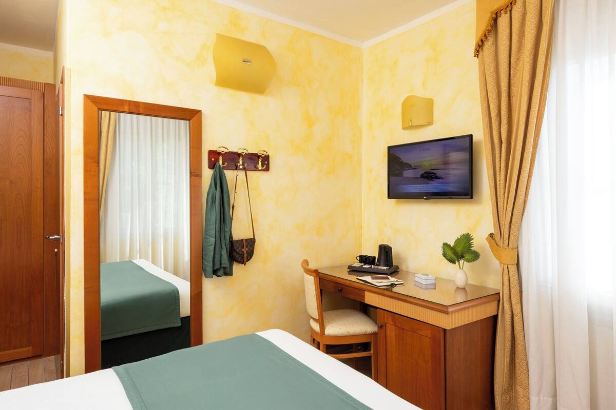 A hotel room with a bed, a desk with a chair, a television, and a window with curtains.