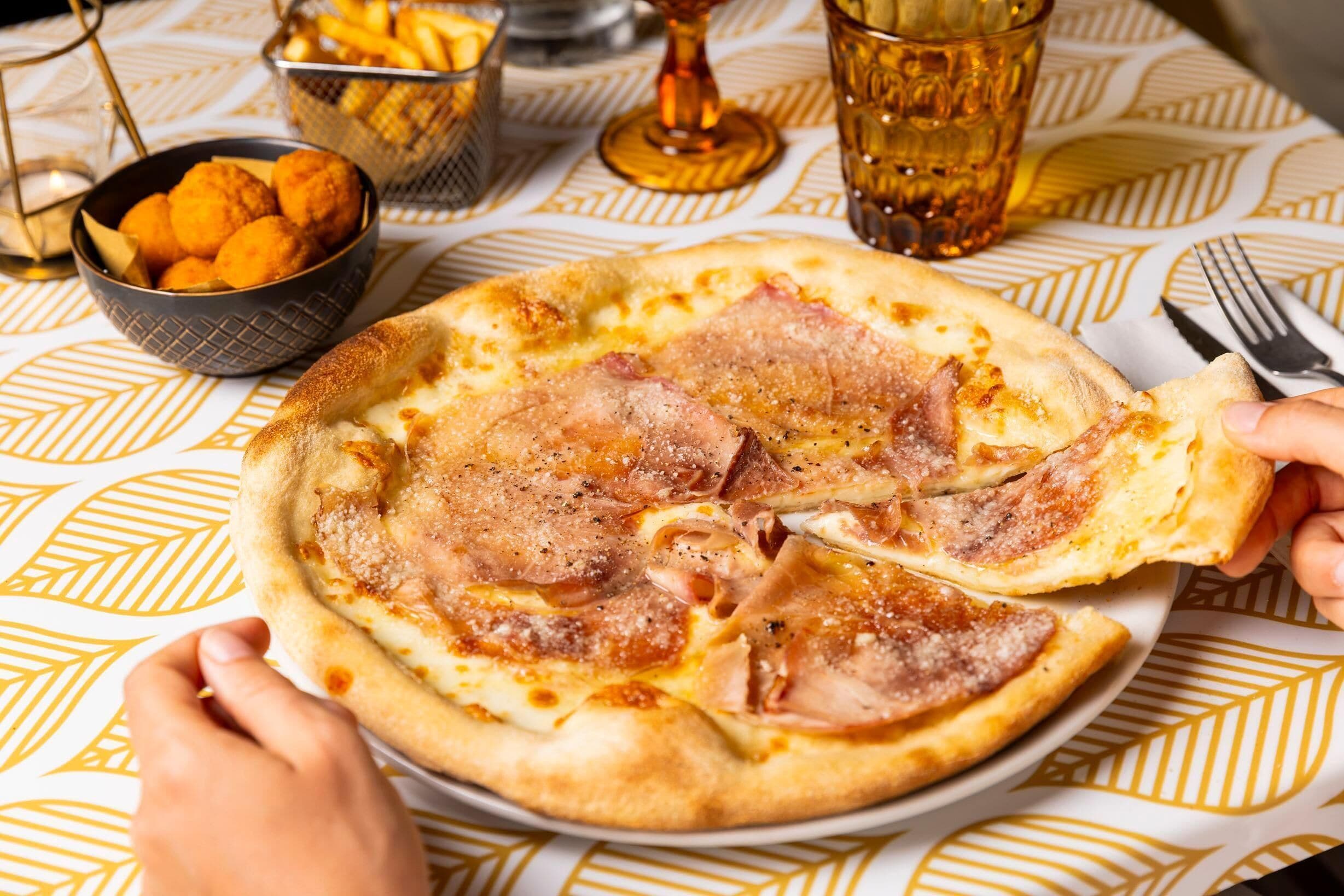Une pizza garnie de jambon, servie sur une assiette, un verre d’un liquide ambré et un panier de frites, le tout posé sur une table recouverte d’une nappe à motifs.