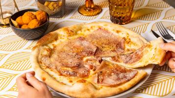Une pizza garnie de jambon, servie sur une assiette, un verre d’un liquide ambré et un panier de frites, le tout posé sur une table recouverte d’une nappe à motifs.