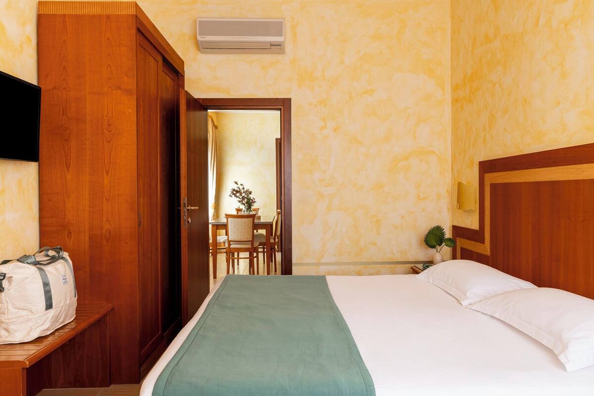 A hotel room with a bed, a wooden wardrobe, a desk with a bag, and a dining area visible through an open door.