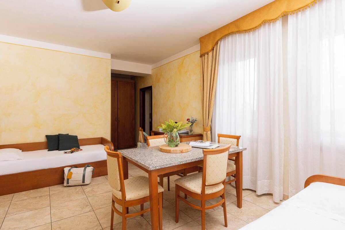 A hotel room with a bed, a dining table with chairs, a sofa, and a window with curtains.