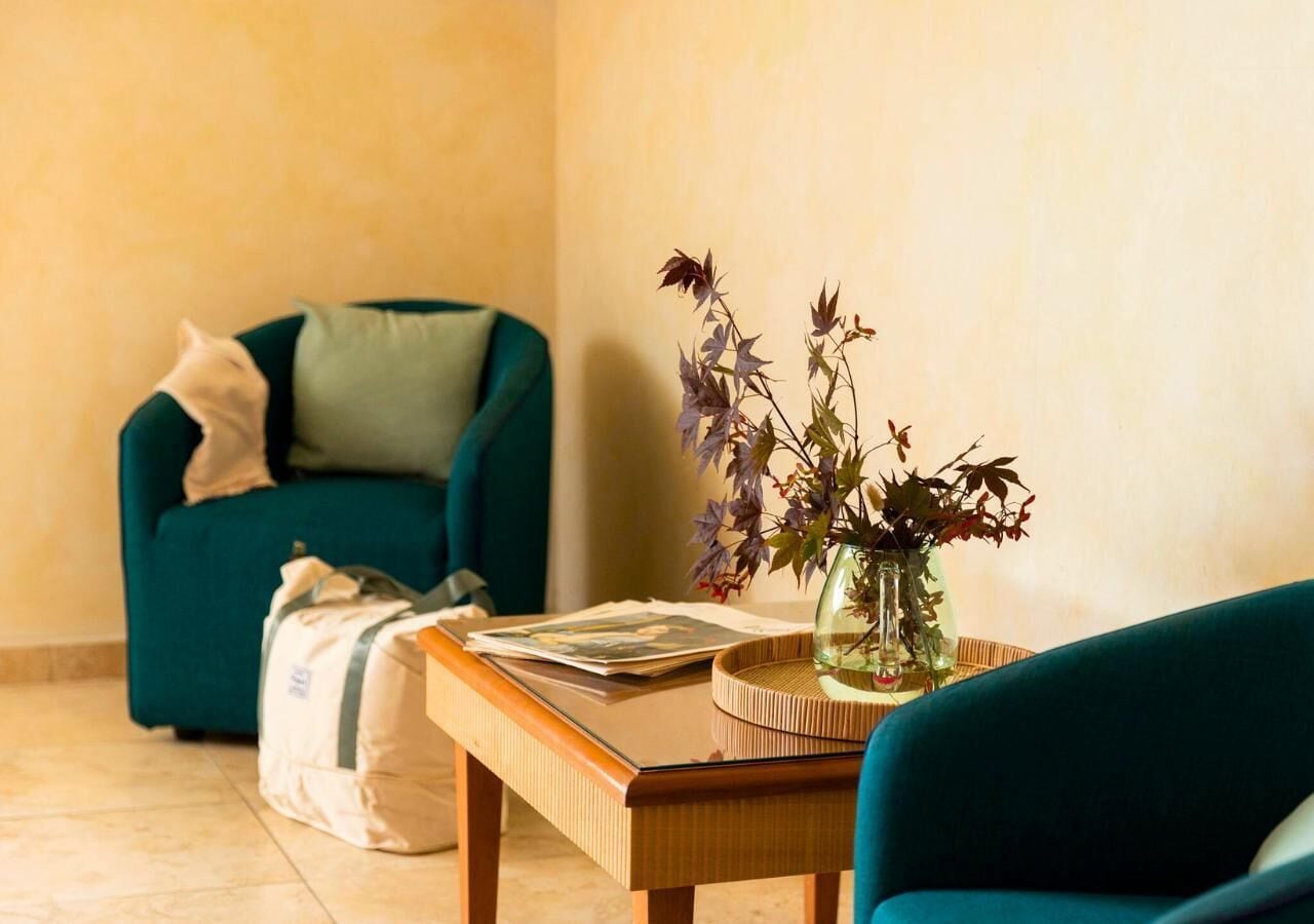 Una stanza con una poltrona color petrolio, un tavolo di legno con un vaso di fiori e una borsa a terra.
