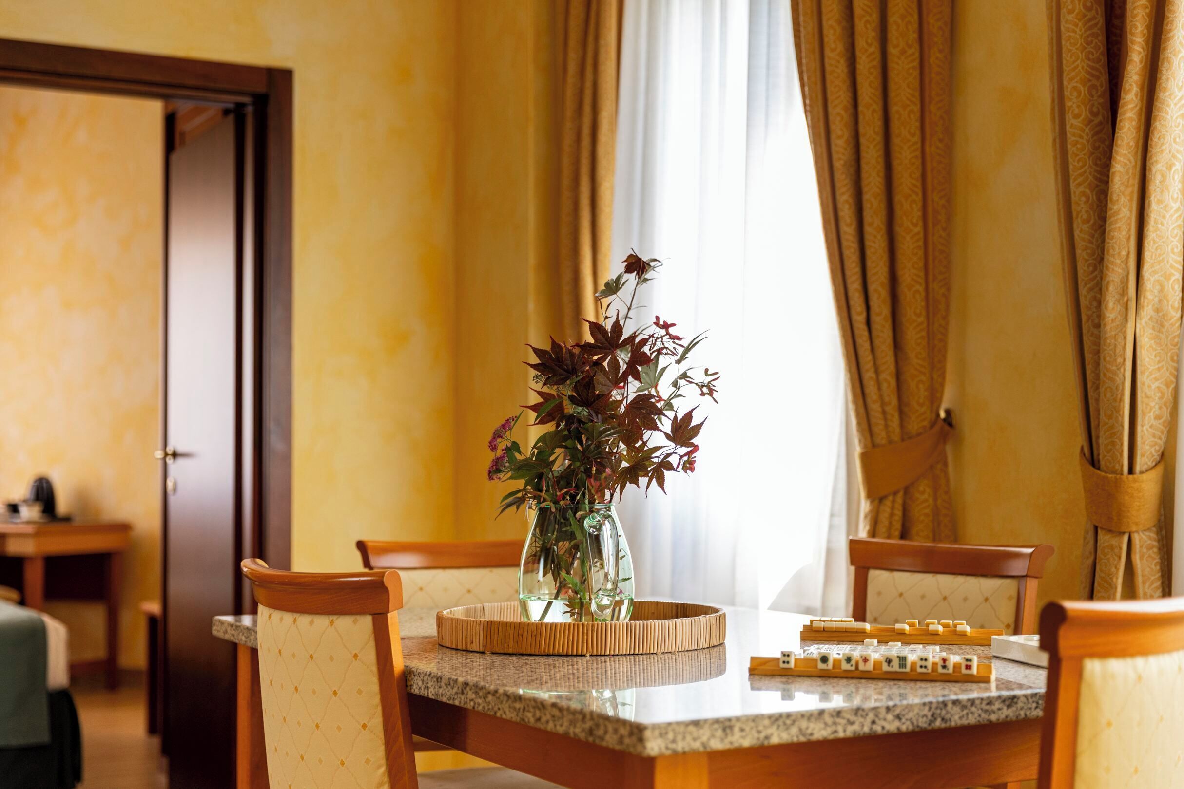 A dining area with a wooden table, chairs, a vase with flowers, and a window with curtains.