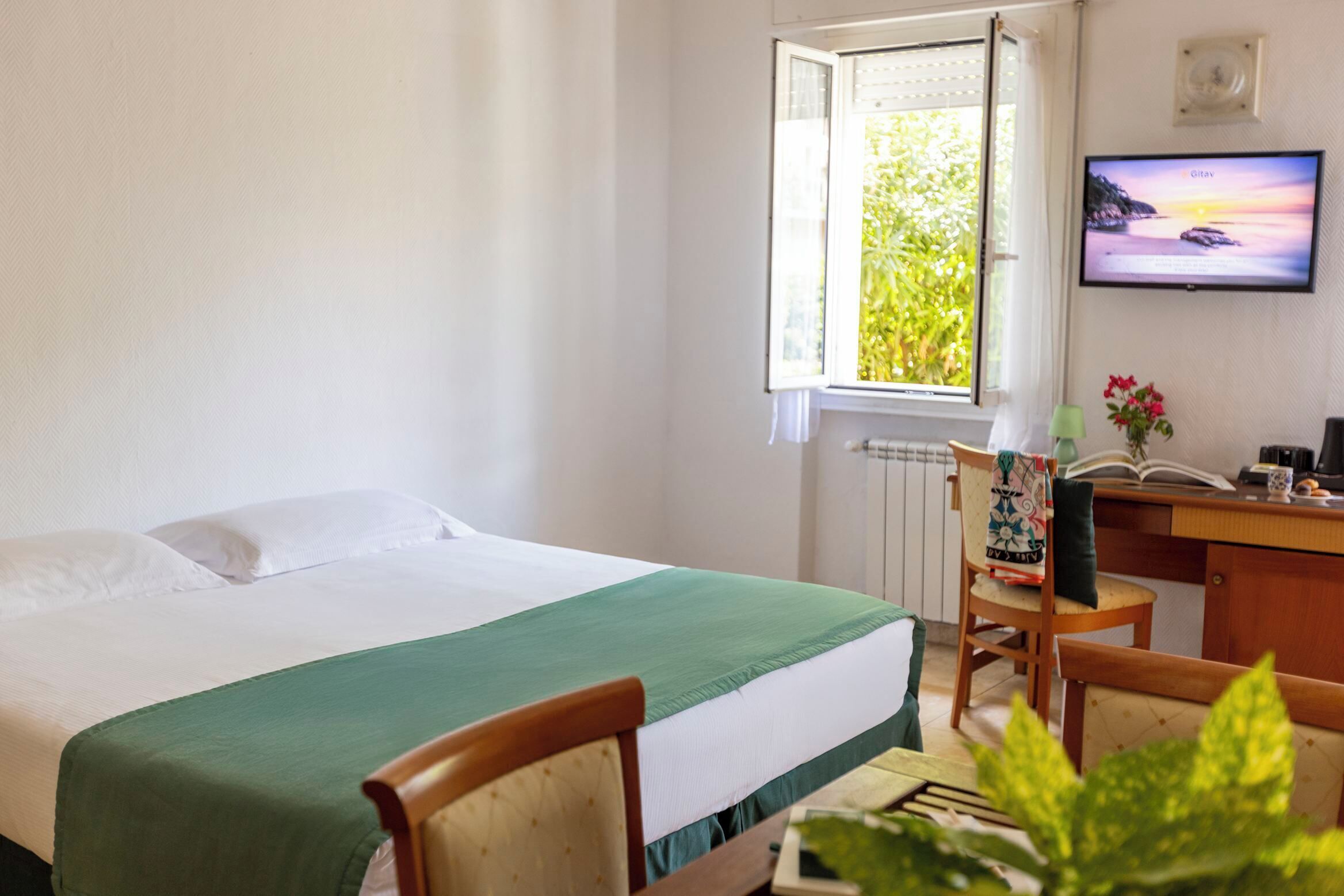Quarto de hotel com cama, escrivaninha e cadeira, televisão e janela com vista para área verde.
