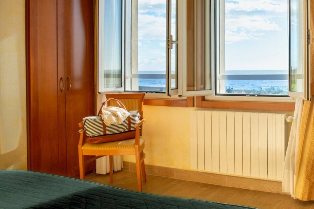 A room with a wooden wardrobe, a chair with a striped bag, a radiator, and a window with a view of the sea.