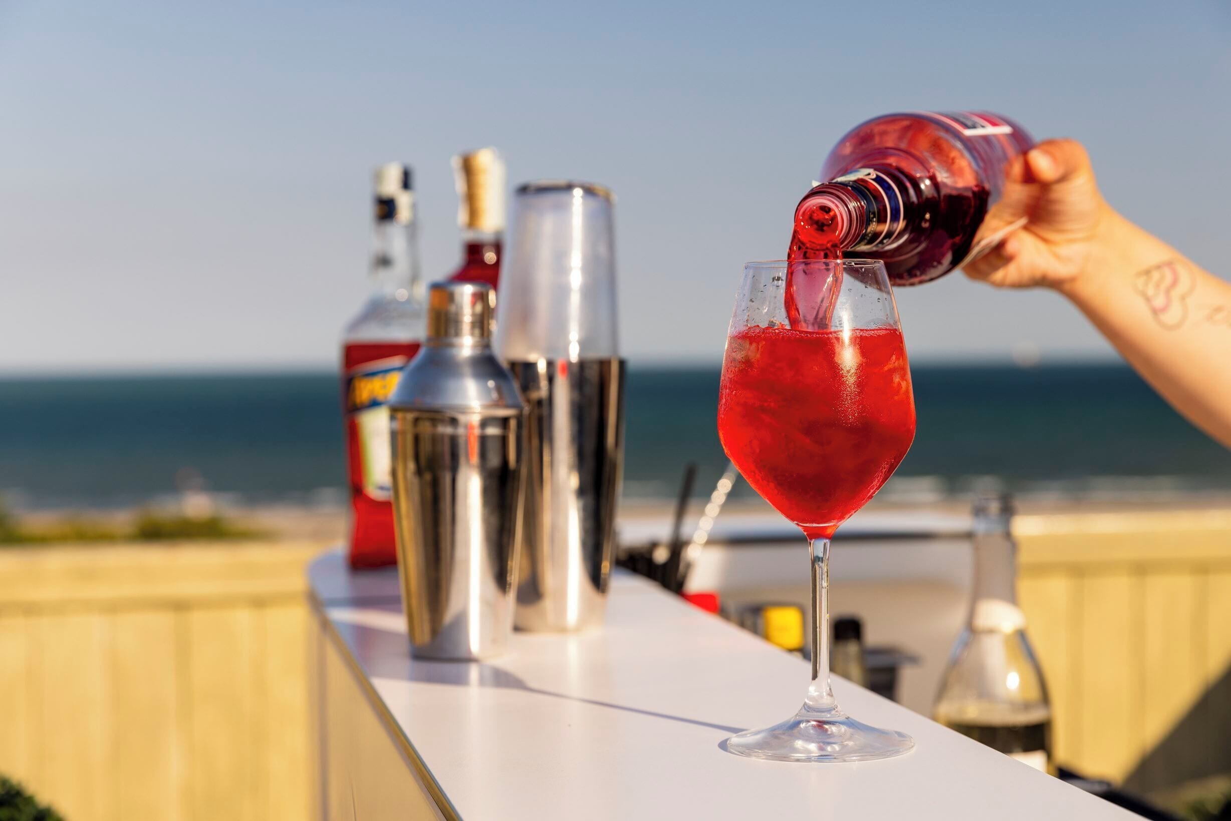 A cocktail being poured into a glass on a bar counter with a view of the ocean in the background.