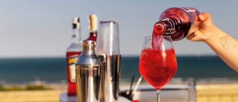 A cocktail being poured into a glass on a bar counter with a view of the ocean in the background.