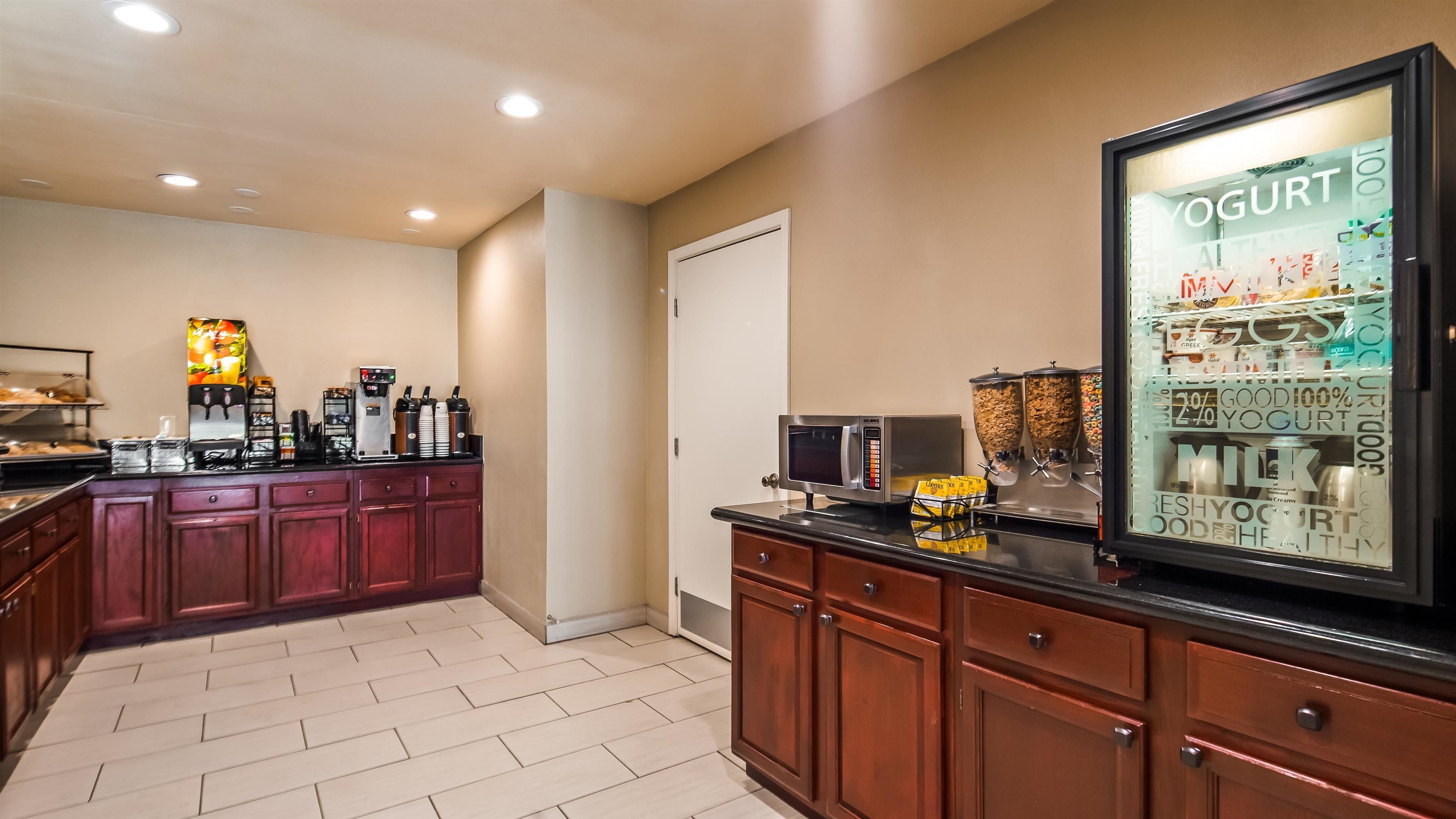 Una cocina de hotel con estación de café, dispensadores de yogur y leche, y un refrigerador que exhibe yogur.