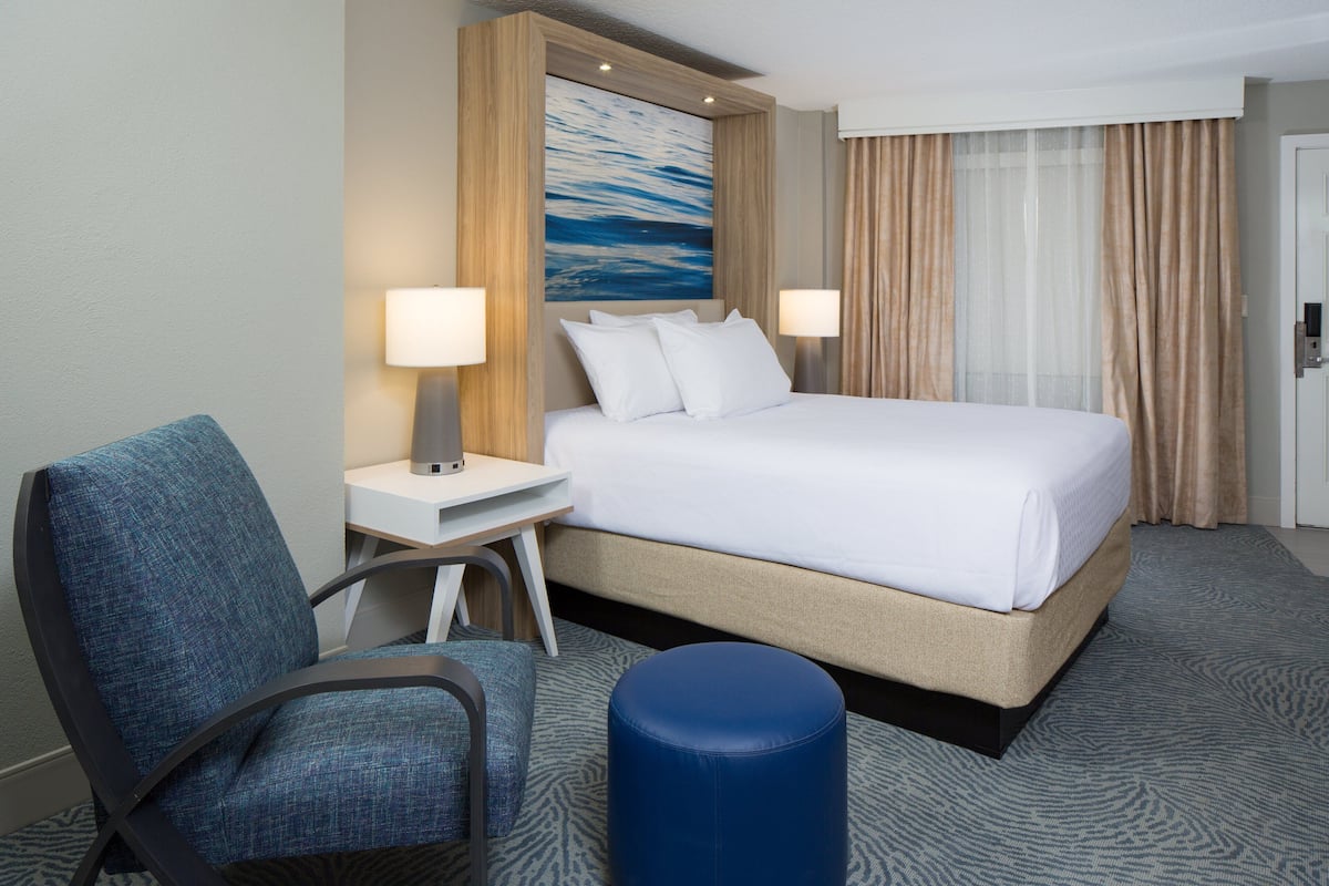 A hotel room with a bed, two bedside lamps, a desk, a chair, and a large window with curtains.