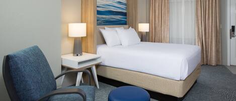 A hotel room with a bed, two bedside lamps, a chair, a desk, a blue ottoman, and a painting on the wall.