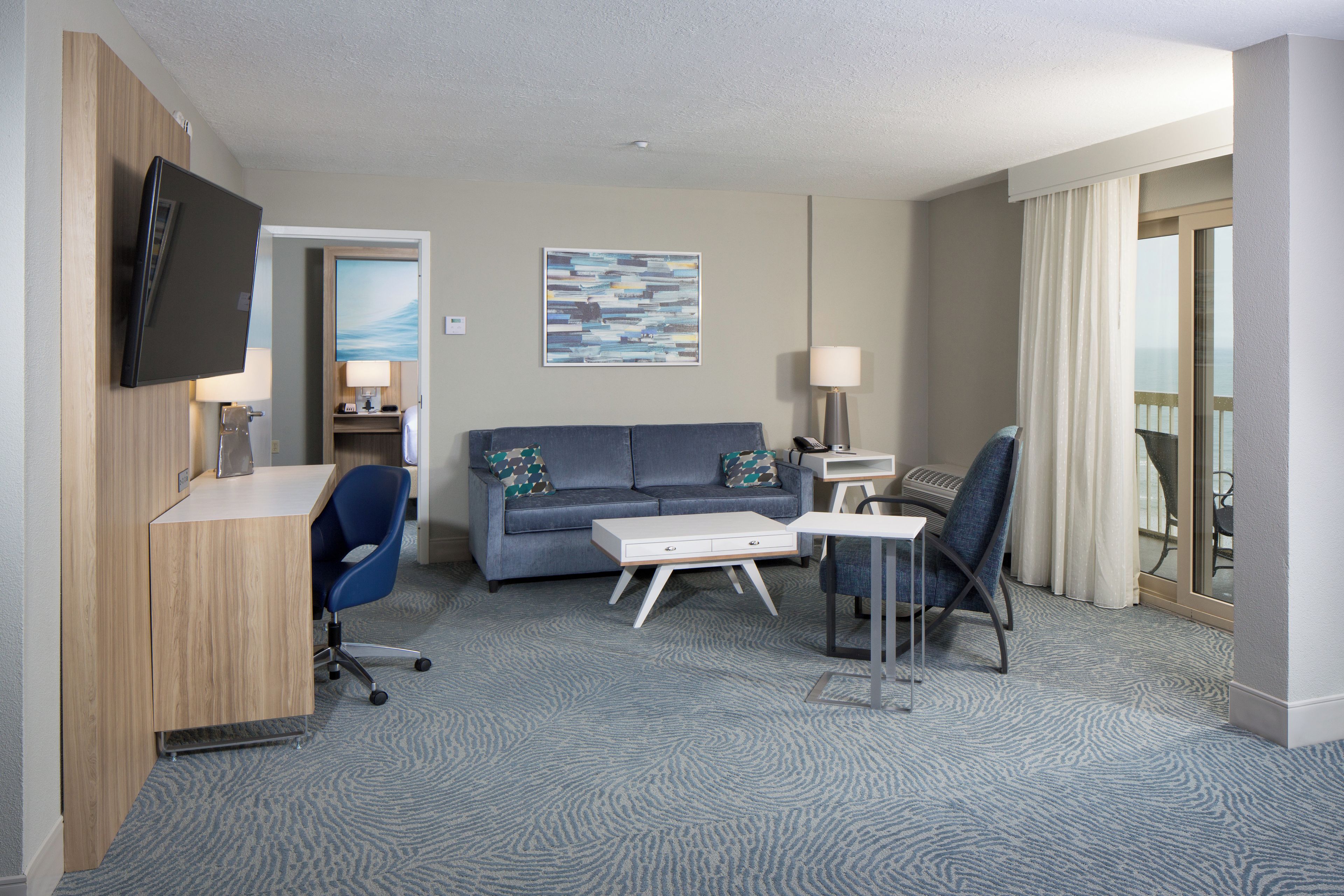 A hotel room with a flat-screen TV mounted on the wall, a sofa with patterned cushions, a desk with a chair, a small table with a lamp, and a view of the sea through the window.