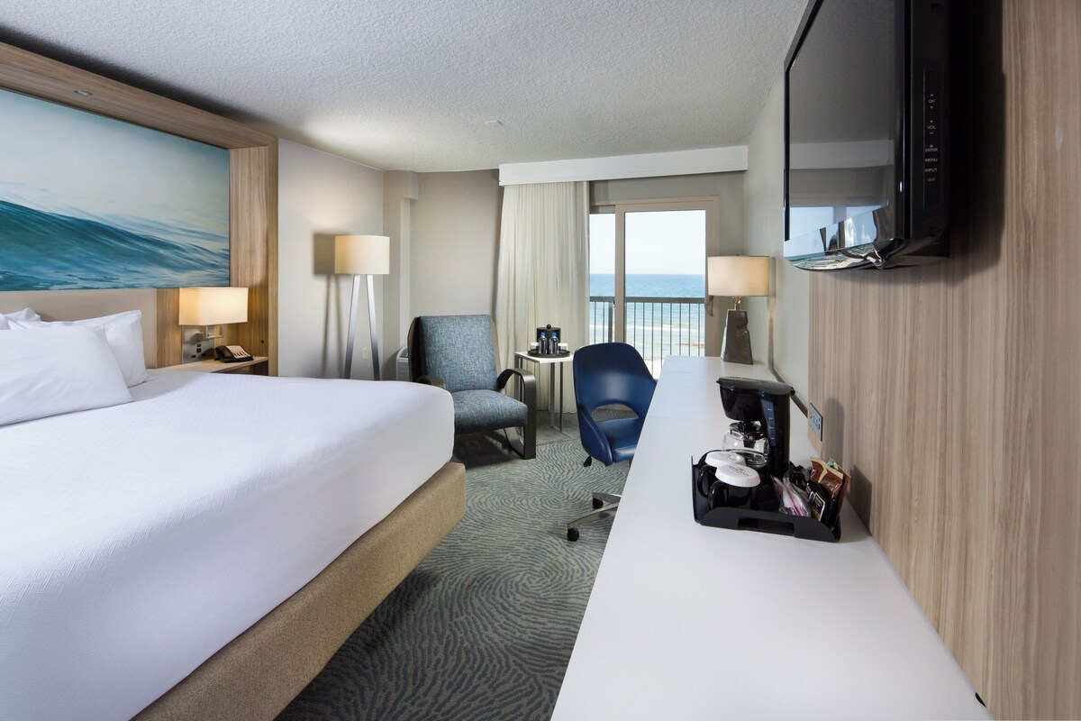 A hotel room with a bed, a desk, a chair, a TV, and a view of the sea.