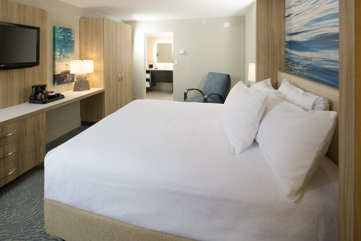 A hotel room with a large bed, a desk with a lamp, a chair, and a TV.